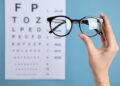 Woman holding glasses against eye chart on blue background, closeup. Ophthalmologist prescription