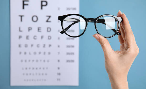 Woman holding glasses against eye chart on blue background, closeup. Ophthalmologist prescription