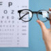 Woman holding glasses against eye chart on blue background, closeup. Ophthalmologist prescription